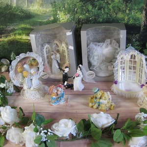 May include: A set of four wedding cake toppers in their original packaging. The toppers feature a bride and groom, a couple of teddy bears, and a pair of figurines with a mirror. The toppers are all white and have a delicate lace design.