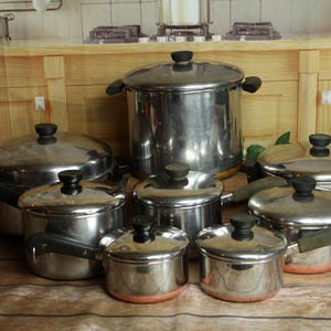 May include: A collection of stainless steel cookware, including pots and pans of various sizes. The pots have black handles and lids with black knobs. The copper-colored bottoms add a touch of warmth to the set.