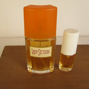 May include: Two perfume bottles on a wooden surface. The larger bottle has an orange cap and a label that reads "LADY STETSON." The smaller bottle has a white cap. Both bottles contain a golden-colored liquid.