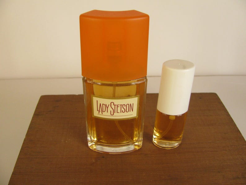 May include: Two perfume bottles on a wooden surface. The larger bottle has an orange cap and a label that reads "LADY STETSON." The smaller bottle has a white cap. Both bottles contain a golden-colored liquid.
