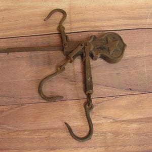 May include: A vintage, rusty, metal hanging scale with two hooks and a star-shaped design on the top. The scale is made of iron and has a rustic look.