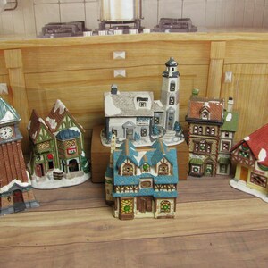 May include: A collection of miniature ceramic village buildings, including a clock tower, houses, and a lighthouse. The buildings feature detailed designs with snow-covered roofs and colorful facades. The scene is set on a wooden surface.