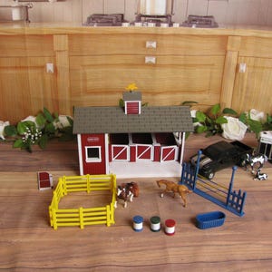 May include: A toy farm set featuring a red and white barn with a gray roof, a yellow fence, and miniature animals. Includes a black truck, horse trailer, and blue and white accessories. The set is on a wooden surface.