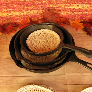 May include: Three cast iron skillets stacked on top of each other. The skillets are on a wooden surface. The top skillet has a crocheted pot holder inside.