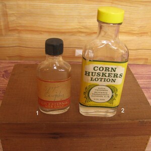 May include: Two clear glass bottles. The smaller bottle on the left has a black cap and a tan label with red text reading FOR EXTERNAL USE ONLY. The larger bottle on the right has a yellow cap and a yellow label reading CORN HUSKERS LOTION.