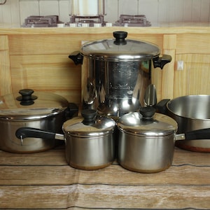 May include: A set of stainless steel pots and pans with black handles and lids. The cookware includes a large stockpot, a saucepan, and several smaller pots, all with a shiny, metallic finish. The pots are arranged on a wooden surface.