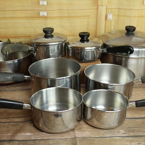 May include: A collection of stainless steel cookware including various pots and pans with black handles. The set features multiple sizes of saucepans and stockpots, some with matching lids, all displaying a polished metallic finish and black knobs.