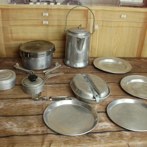 May include: A collection of vintage camping cookware, including a kettle with a handle, a pot, a small stove, a mess kit, and several plates. The items are made of metal and are arranged on a wooden surface.