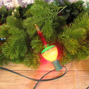 May include: A vintage Christmas light bulb with a green and red glass bulb and a yellow light inside. The bulb is surrounded by green pine branches.