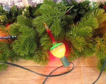 May include: A vintage Christmas light bulb with a green and red glass bulb and a yellow light inside. The bulb is surrounded by green pine branches.