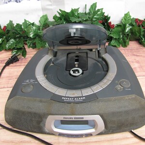 May include: A gray Philips CD clock radio with a digital display and a CD player. The CD player is open and the CD tray is visible. The radio has a repeat alarm and brightness control.