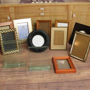 May include: A collection of various picture frames in different styles and colors. Frames include gold, black, brown, and clear glass. Some frames are rectangular, while others are round. The frames are arranged on a wooden surface.
