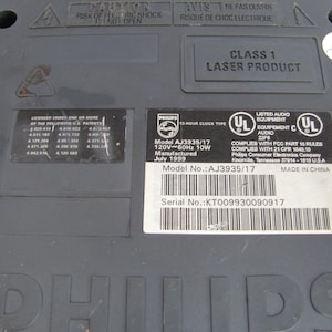 May include: Close-up of a black Philips laser disc player with a model number AJ3935/17 and serial number KT009930090917. The player is labeled as Class 1 Laser Product and has a 12-hour clock type. It is made in China and manufactured in July 1999.
