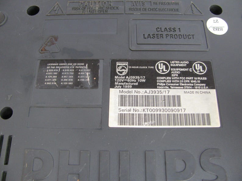 May include: Close-up of a black Philips laser disc player with a model number AJ3935/17 and serial number KT009930090917. The player is labeled as Class 1 Laser Product and has a 12-hour clock type. It is made in China and manufactured in July 1999.