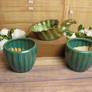 May include: A collection of ceramic pottery in shades of green and brown. The set includes two small, ribbed teal bowls, and a larger, shell-shaped dish. The pottery is displayed on a wooden surface with white flowers in the background.