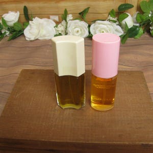 May include: Two perfume bottles with amber liquid. One has a cream-colored, geometric cap, the other a pink cap. They sit on a wooden block, with white roses and greenery in the background. The bottles are likely vintage.