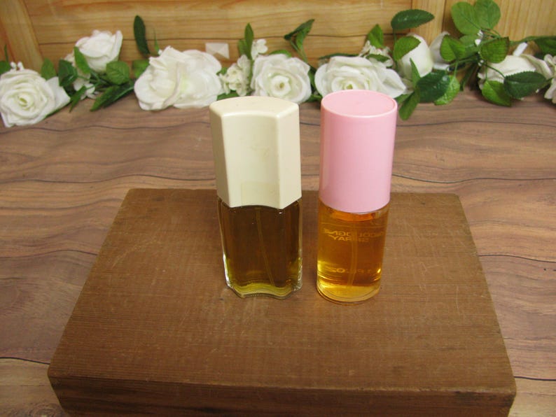 May include: Two perfume bottles with amber liquid. One has a cream-colored, geometric cap, the other a pink cap. They sit on a wooden block, with white roses and greenery in the background. The bottles are likely vintage.