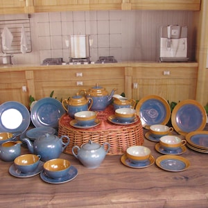 May include: A blue and gold tea set with a teapot, creamer, sugar bowl, cups, and saucers. The set is arranged on a wicker table with a floral background.