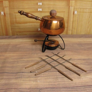 May include: A copper fondue pot with a wooden handle and lid, set on a black metal stand. Six fondue forks with wooden handles are arranged in front of the pot. The pot is approximately 6 inches in diameter.