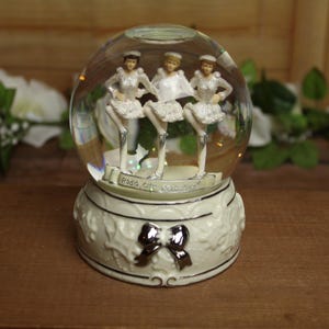 May include: A snow globe featuring three figurines of dancers in white costumes and hats. The globe sits on a white base with a silver bow. The text "Radio City Rockettes" is visible.