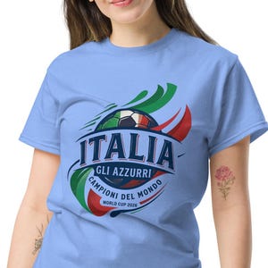 Puede incluir: Una camiseta azul claro con la palabra "ITALIA" y un diseño de balón de fútbol con los colores de la bandera italiana. La camiseta también incluye el texto "GLI AZZURRI! CAMPIONI DEL MONDO WORLD CUP 2026".