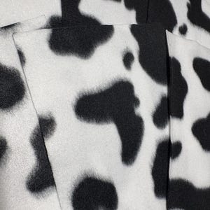 May include: Close-up of a fabric with a cow print pattern. The fabric is white with irregular black spots. The texture appears soft and possibly fleece-like, with visible fibers.