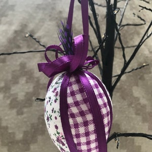 May include: Purple and white fabric Easter egg with a purple ribbon and a black feather hanging from a branch.