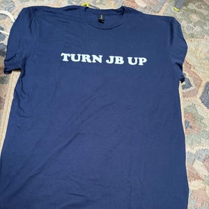 May include: Navy blue t-shirt with the text "TURN JB UP" in white block letters. The shirt is laid flat on a patterned surface. The t-shirt has short sleeves and a crew neck.
