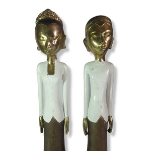 Peut inclure: Deux statues de personnes en vêtements traditionnels peintes en or et en blanc. Les statues sont debout sur des bases carrées.