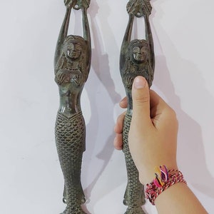 May include: Two bronze mermaid door pulls. Each mermaid figure holds a star above her head. The mermaid's tail is a fin. The door pulls are a dark bronze color and are mounted on a white wall.
