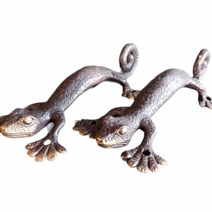 May include: Two bronze-colored lizard-shaped door handles. The handles are detailed with scales and have curled tails. The lizards are in a crawling position, with their front and back legs extended. The handles are designed to be mounted on a door or drawer.