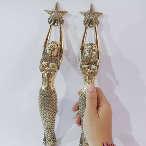 May include: Two brass mermaid-shaped door handles. Each mermaid holds a starfish above her head. The handles have detailed scales and flowing hair. The mermaids are gold-colored and stand against a white background.