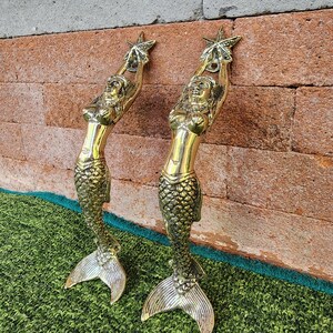 May include: Two gold-colored mermaid sculptures, each holding a star above their heads. The mermaids have detailed scales and flowing hair. The sculptures are displayed against a brick wall and green surface.