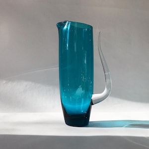 May include: A teal blue glass jug with a curved handle. The jug has a rounded base and a wide mouth.