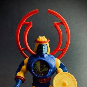 Puede incluir: Una figura de acción azul y amarilla con un casco rojo y blanco y un escudo circular rojo. La figura sostiene un escudo amarillo con un diseño circular.