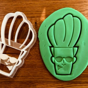 Puede incluir: Un cortador de galletas de plástico blanco con forma de personaje de dibujos animados con una versión de masa de galletas esculpida en verde del mismo personaje a su lado. El personaje tiene un gran peinado verde puntiagudo, gafas de sol y un pequeño bigote puntiagudo.