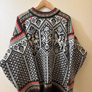 May include: A long-sleeve crew neck sweater featuring a complex geometric and animal-inspired pattern in black, white, red, and green. The design includes repeating hexagonal motifs, intricate borders, and a small rectangular patch on the lower sleeve.