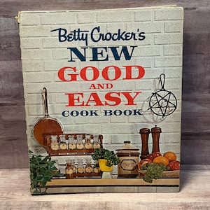 May include: A vintage Betty Crocker cookbook titled "New Good and Easy Cookbook". The cover features a brick wall background with a kitchen scene including a spice rack, a cookie jar, and a bowl of fruit.