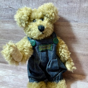 May include: A small, tan teddy bear wearing blue denim overalls with a green patch that reads "Only's Bears Tim Gearwear".