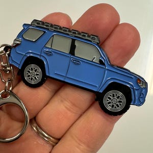 Puede incluir: Un llavero de metal azul con forma de Toyota 4Runner con baca. El llavero tiene un anillo plateado para sujetarlo a las llaves.