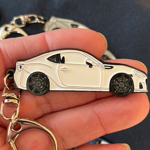 May include: White metal keychain featuring a detailed model of a Subaru BRZ sports car. The keychain has a silver split ring and a black metal loop.