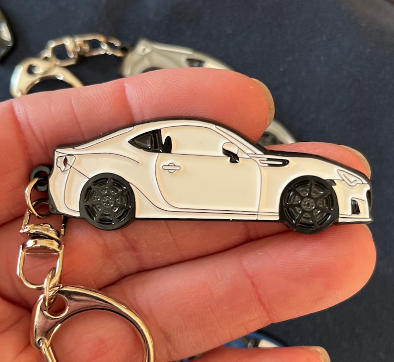 May include: White metal keychain featuring a detailed model of a Subaru BRZ sports car. The keychain has a silver split ring and a black metal loop.