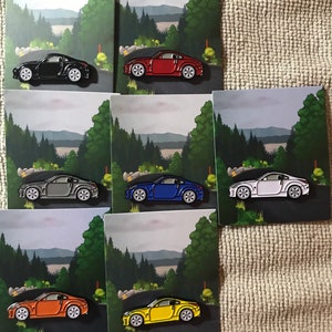 May include: Set of six enamel pins featuring a cartoon illustration of a Nissan 350Z sports car in six different colors: black, red, gray, blue, white, and yellow. Each pin is set against a background of a winding road through a forest.