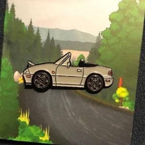 Puede incluir: Un pin de esmalte plateado de un coche deportivo convertible con ruedas negras y un interior negro. El coche está conduciendo por un camino sinuoso con árboles verdes y una cordillera en el fondo.