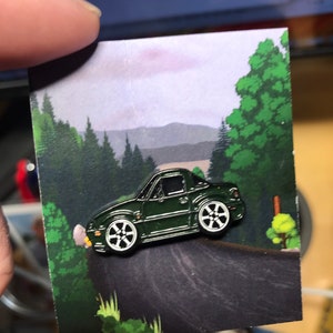 Puede incluir: Pin de esmalte con forma de coche verde oscuro con ruedas blancas. El pin se muestra en una tarjeta con una ilustración de una carretera que serpentea por un bosque. El pin está sujeto por los dedos de una persona.