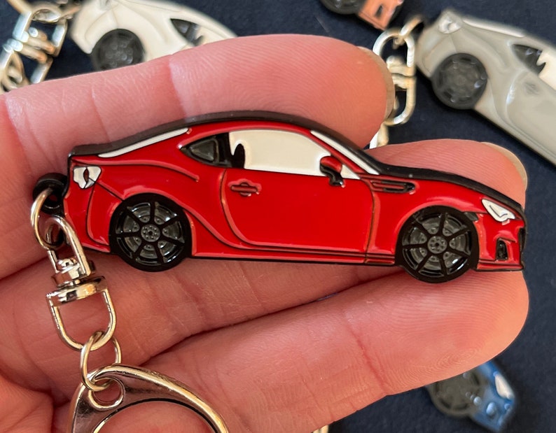 May include: A red metal keychain shaped like a sports car with a white interior and black wheels. The keychain has a silver metal ring.