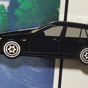 May include: Black enamel pin of a black station wagon with white star-shaped wheels. The pin is on a white background.