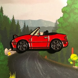 Puede incluir: Pin de coche descapotable rojo con ruedas negras e interior blanco. El pin está sobre un fondo de carretera que serpentea por un bosque, con una combinación de colores verde y azul.