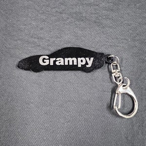 Puede incluir: Llavero negro con forma de coche con la palabra "Grampy" en letras blancas. El llavero tiene un cierre y un anillo plateados para sujetarlo a llaves o a un bolso. El fondo es de tela gris.