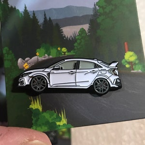Könnte beinhalten: Ein weißer Emaille-Pin mit einer weißen Honda Civic Type R mit schwarzen Akzenten. Das Auto ist im Profil dargestellt, wobei die Fahrerseite zum Betrachter zeigt. Der Pin ist vor dem Hintergrund einer Cartoon-Landschaft mit Bäumen und einer kurvigen Straße platziert.